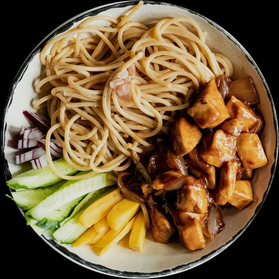A bowl of noodles with glazed chicken, cucumber, peppers and red onion